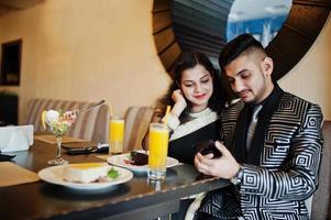 encantadora pareja india enamorada, vestida con sari y traje elegante, sentada en el restaurante y mirando el teléfono juntos. en la mesa pasteles de postre, helados y jugos. foto