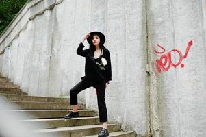 Sensual girl all in black, red lips and hat. Goth dramatic woman hold white chrysanthemum flower against graffiti wall. photo