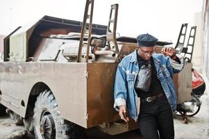 African american man in jeans jacket, beret and eyeglasses, smoking cigar and posed against btr military armored vehicle. photo