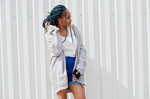 African woman with dreads hair, in jeans shorts  posed against white steel wall with mobile phone in hand. photo