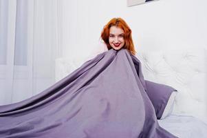 Portrait of an attractive redheaded girl laying on the bed and covering herself with a purple blanket. photo
