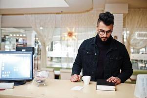 Arab man wear on black jeans jacket and eyeglasses in cafe drink coffee at bar with book. Stylish and fashionable arabian model guy. photo