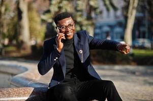 Amazingly looking african american man wear at blue blazer with brooch, black turtleneck and glasses posed at street. Fashionable black guy speaking on phone and show something. photo