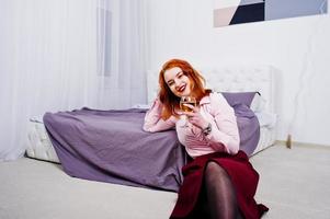 Gorgeous red haired girl in pink blouse and red skirt with glass of wine at hand near bed at room. photo