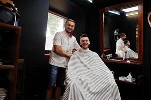 Young bearded man getting haircut by hairdresser while sitting in chair at barbershop. Barber soul. photo