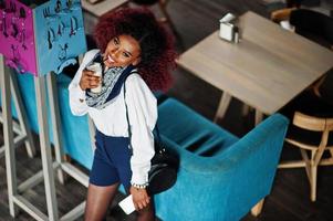 Attractive african american curly girl in white blouse and blue shorts posed at cafe with latte and mobile phone at hand. View from above. photo