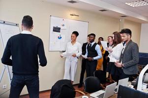 Mixed race business coach presenting report standing near whiteboard pointing on sales statistic shown on diagram and chart teach diverse company members gathered together in conference room. photo