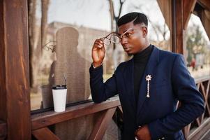 Amazingly looking african american man wear at blue blazer with brooch, black turtleneck and glasses posed at street. Fashionable black guy with cup of coffe. photo