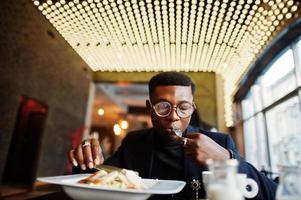 hombre afroamericano de moda con traje y gafas sentado en el café y comiendo ensalada. foto