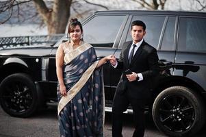 Elegant and fashionable indian friends couple of woman in saree and man in suit posed against rich black suv car. photo