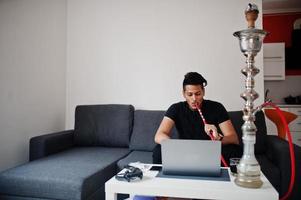 Handsome and fashionable indian man in black sitting at room, smoking hookah and working on laptop. photo