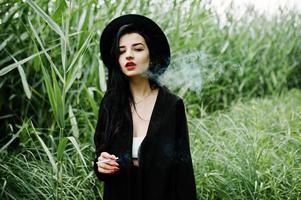 Sensual smoker girl all in black, red lips and hat. Goth dramatic woman smoking on common reed. photo