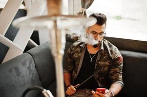 Stylish beard arabian man in glasses and military jacket smoking hookah at street bar. Arab model having rest and looking on phone. photo