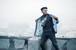 hombre afroamericano con chaqueta de jeans, boina y anteojos posados en un techo abandonado. foto