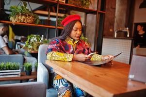 Enthusiastic african american woman in trendy coloured outfit with red beret chilling in cozy cafe, reading menu. photo
