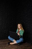 Stylish blonde girl in jacket and jeans with laptop against brick black wall at studio. photo