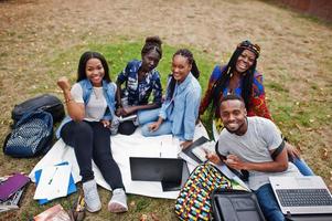 grupo de cinco estudiantes universitarios africanos que pasan tiempo juntos en el campus en el patio de la universidad. amigos afro negros sentados en el césped y estudiando con computadoras portátiles. foto