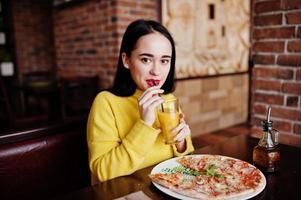 chica morena divertida en suéter amarillo comiendo pizza en el restaurante. foto