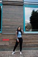 chica afroamericana hipster con camisa de jeans con mangas de leopardo posando en la calle contra una casa de madera con ventanas.