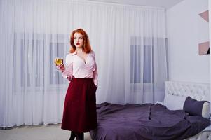 hermosa chica pelirroja con blusa rosa y falda roja con una copa de vino a mano cerca de la cama en la habitación. foto