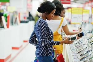 Beautiful three well-dressed afro american girls customers with colored shopping bags in mobile phone shop choosing smartphone. photo