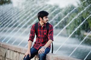 Young indian student man at checkered shirt and jeans with backpack posed on evening city against fountains. photo