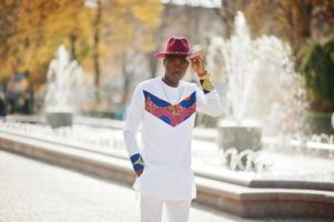 Stylish trendy afro france man in red hat and white outfit posed at autumn day. Black african model guy against fountain. photo