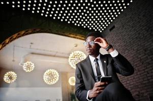 Business african american man wear on black suit and glasses at office looking on mobile phone against futuristic lights. photo