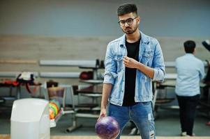 Stylish asian man in jeans jacket and glasses standing at bowling alley with ball at hand. photo