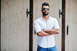 Stylish tall arabian man model in white shirt, jeans and sunglasses posed at street of city. Beard attractive arab guy. photo