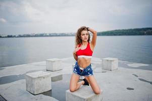 Sexy curly model girl in red top, jeans denim shorts and sneakers posed at stone cubes against cloudy sky and lake. photo