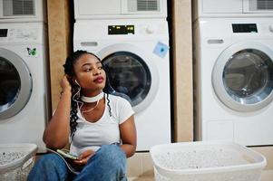 alegre mujer afroamericana sentada con auriculares y leyendo una revista cerca de la lavadora en la lavandería de autoservicio. foto