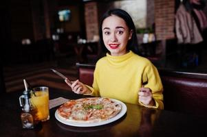 chica morena divertida en suéter amarillo comiendo pizza en el restaurante. foto