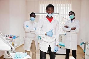 Three african american male doctor with crossed arms in dental clinic. Show thumb up. photo