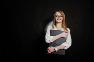 Studio portrait of blonde businesswoman in glasses, white blouse and black skirt holding laptop against dark background. Successful woman and stylish girl concept. photo