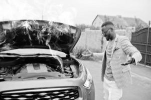 Stylish and rich african american man stand in front of a broken suv car needs assistance looking under opened hood with smoke. photo