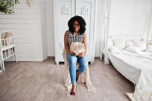 Beautiful african american woman with curly afro hair and eyeglasses posed in room, sitting on retro chair with decorative rose in hands. photo