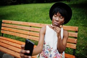 Increíble mujer modelo afroamericana con pantalones verdes y sombrero negro posada en un banco en el parque y haciendo selfie en el teléfono móvil. foto