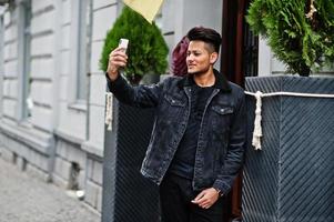 Handsome and fashionable indian man in black jeans jacket posed outdoor and making selfie on phone. photo