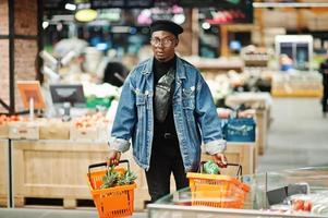 elegante hombre afroamericano casual con chaqueta de jeans y boina negra sosteniendo dos canastas, caminando y comprando en el supermercado. foto