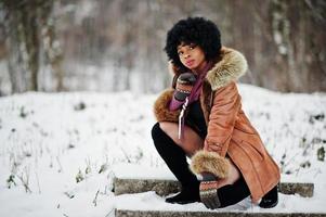 mujer afroamericana de pelo rizado con abrigo de piel de oveja y guantes posados en el día de invierno. foto