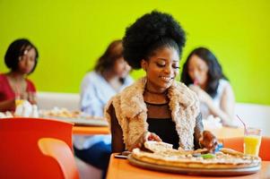 African woman with pizza sitting at restaurant against dark skinned girls. photo