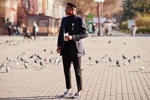 un hombre afroamericano de aspecto asombroso usa un blazer azul con broche, cuello alto negro y anteojos posados en la calle. chico negro de moda con una taza de café. foto