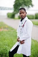 joven médico afroamericano de bata blanca con un estetoscopio posado al aire libre. foto