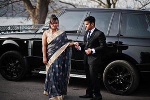 Elegant and fashionable indian friends couple of woman in saree and man in suit posed against rich black suv car. photo