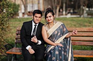 Elegant and fashionable indian friends couple of woman in saree and man in suit sitting on bench. photo