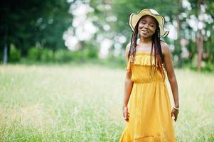 retrato de una hermosa mujer afroamericana de 20 años vestida con vestido amarillo y sombrero de verano posando en la hierba verde en el parque. foto