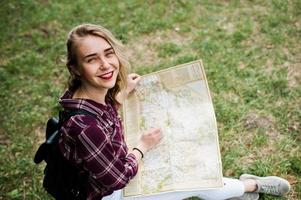 retrato de una joven y hermosa rubia positiva sentada en el suelo con un mapa en las manos en el bosque. foto