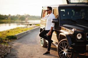 Solid asian man in white shirt and sunglasses posed near black mafia suv car. photo