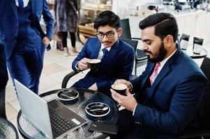 Two indian business man in suits sitting at office on cafe, looking at laptop and drinking coffee. photo